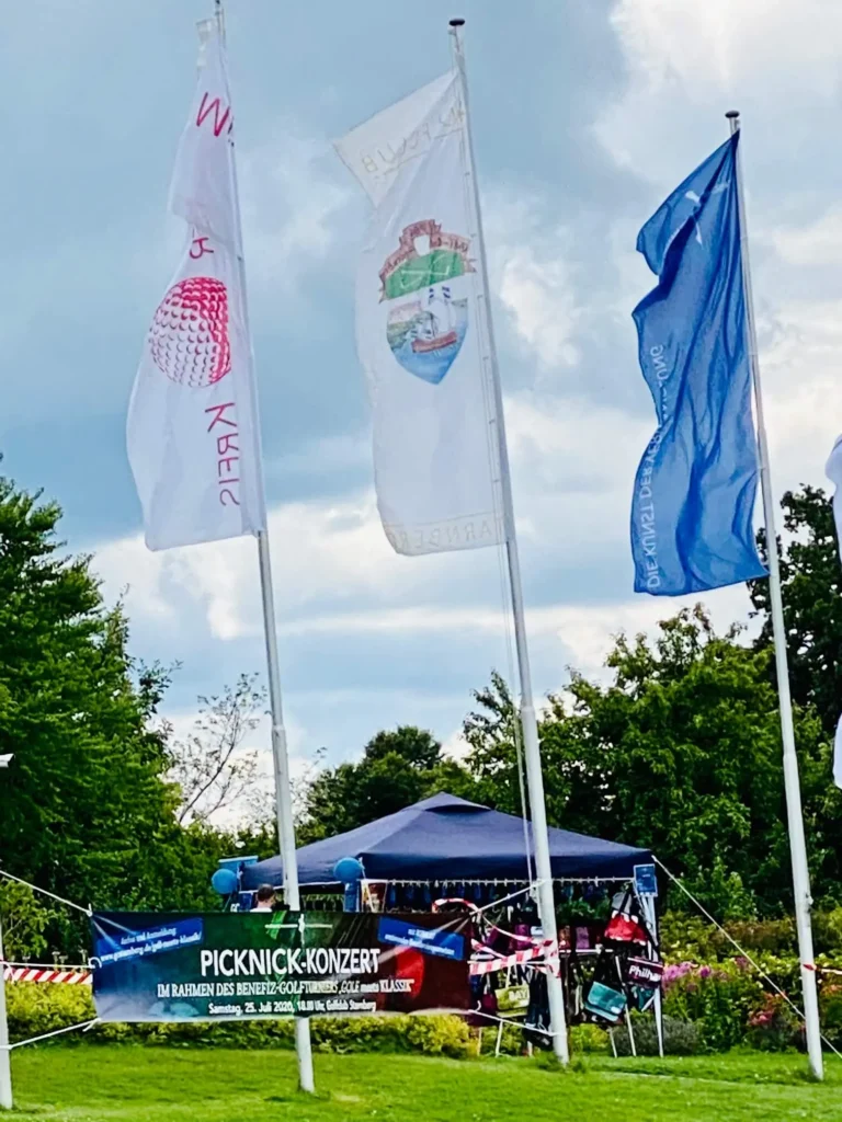 Drei Veranstaltungsflaggen auf einer Wiese vor einem Zelt und Bäumen; darunter ein Banner für ein Picknick-Konzert im Rahmen eines Benefiz-Golfturniers.